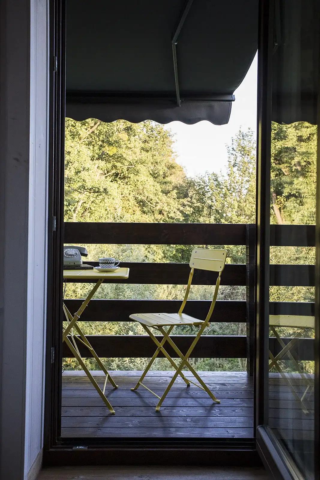 Balkon z markizą widziany przez otwarte drzwi: składany stolik i krzesło, w tle las